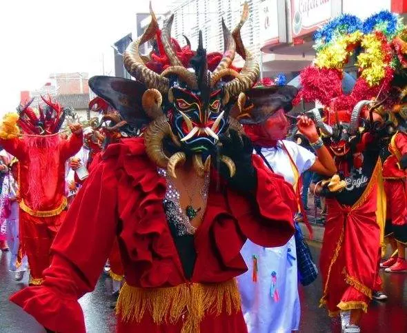 Persona con un traje rojo y una máscara con cuernos participando en un vibrante desfile callejero con otras personas enmascaradas y disfrazadas en el fondo.