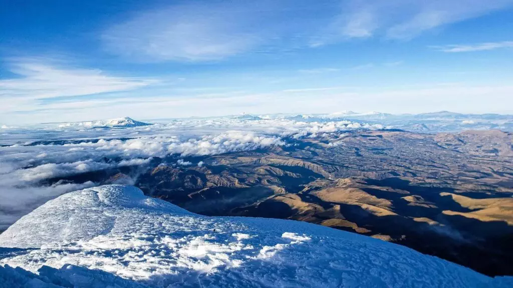 Vista desde un pico nevado con vistas a un vasto paisaje de colinas onduladas y montañas distantes bajo un cielo azul con nubes dispersas.